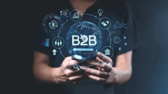 Person holding a smartphone with a glowing digital interface showing the text “B2B” surrounded by icons representing finance, data, supply chain, and business connections.