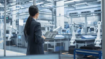 business woman holding laptop, looking out over manufacturing floor focused on manufacturing cost reduction