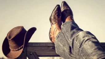 A cowboy's legs and boots resting on a wooden fence, crossed at ankle; his brown cowboy hat is resting on the end of the fence, and he's planning his agenda for devops at opentext world 2025