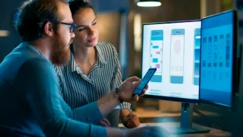 Two developers comparing their mobile screen with what they see on their two desktop monitors, indicating the new features in OpenText Application Quality Management 26.1 release.
