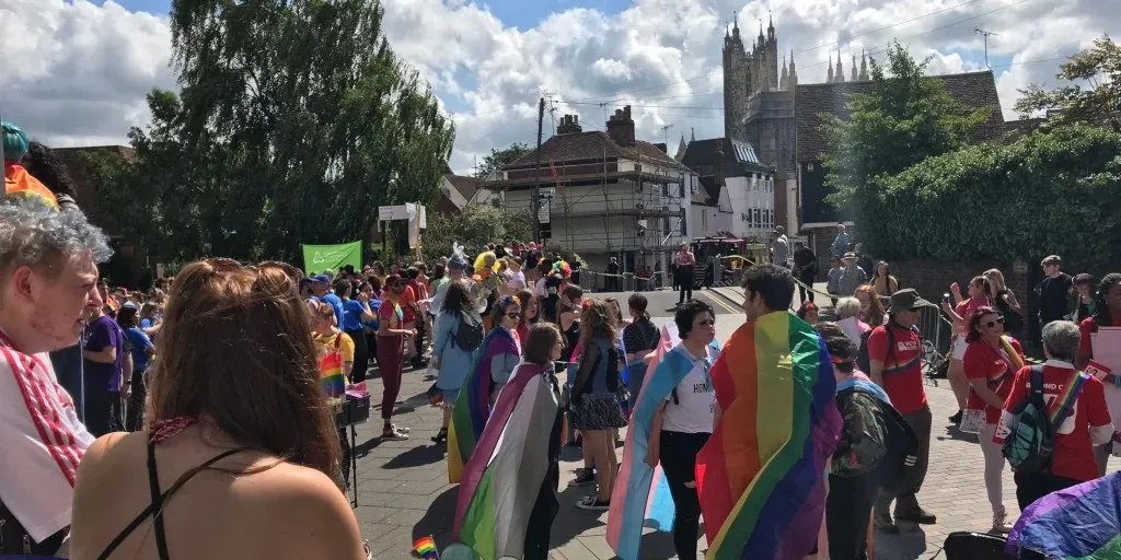 A large group of people stand together waiting for the parade to start. All of them are wearing different kinds of Pride flags: the traditional rainbow, the trans flag, the ace flag, and many others. Far in the distance, the spires of Canterbury Cathedral poke above the city.